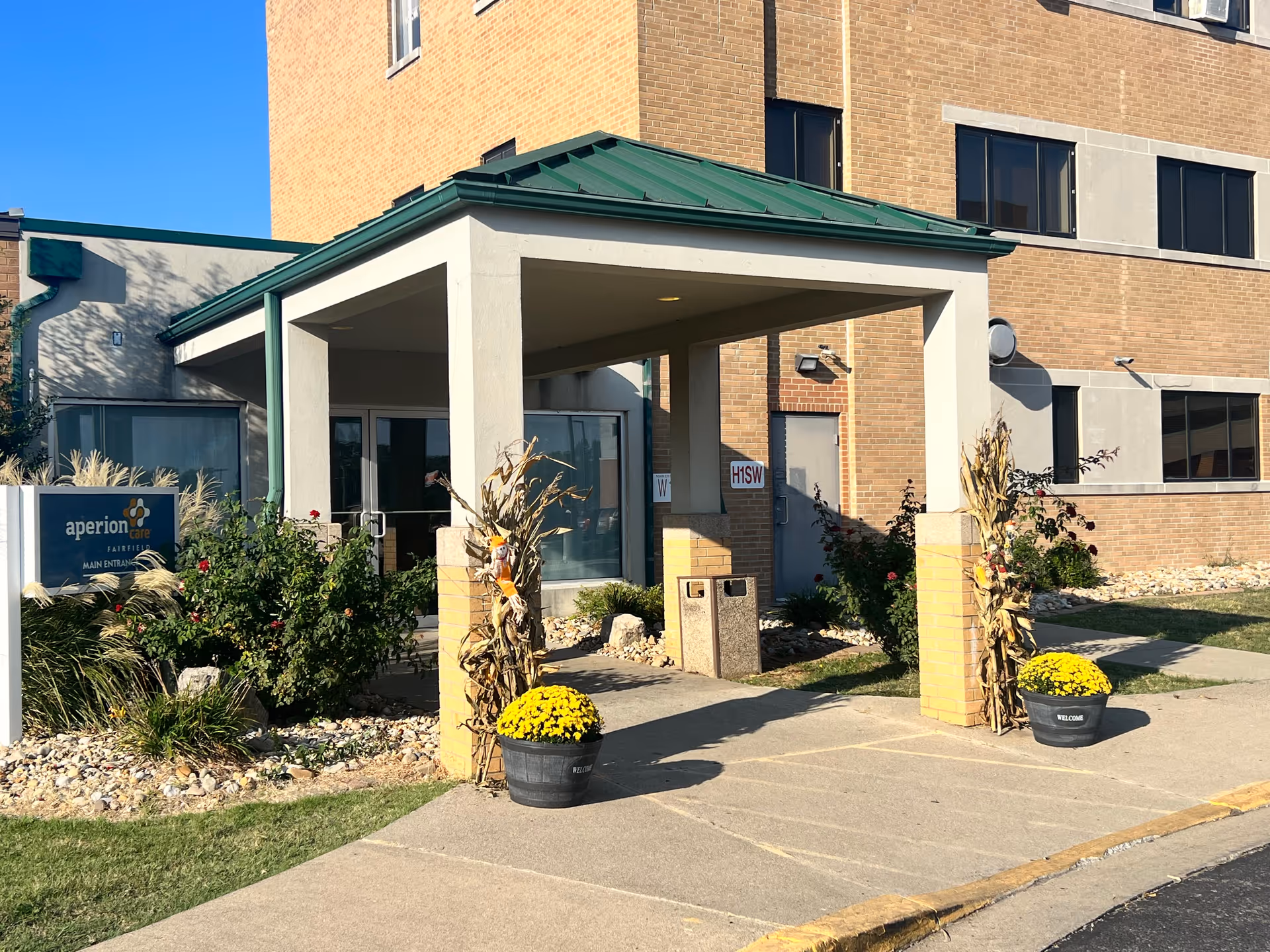 Entrance to a senior living and rehabilitation facility with a covered drop-off area supported by white pillars. There are two black planters with yellow flowers and cornstalk decorations on either side of the entrance. The building is made of brick with several windows, and a sign near the entrance reads 'aperion care FAIRFIELD MAIN ENTRANCE'.