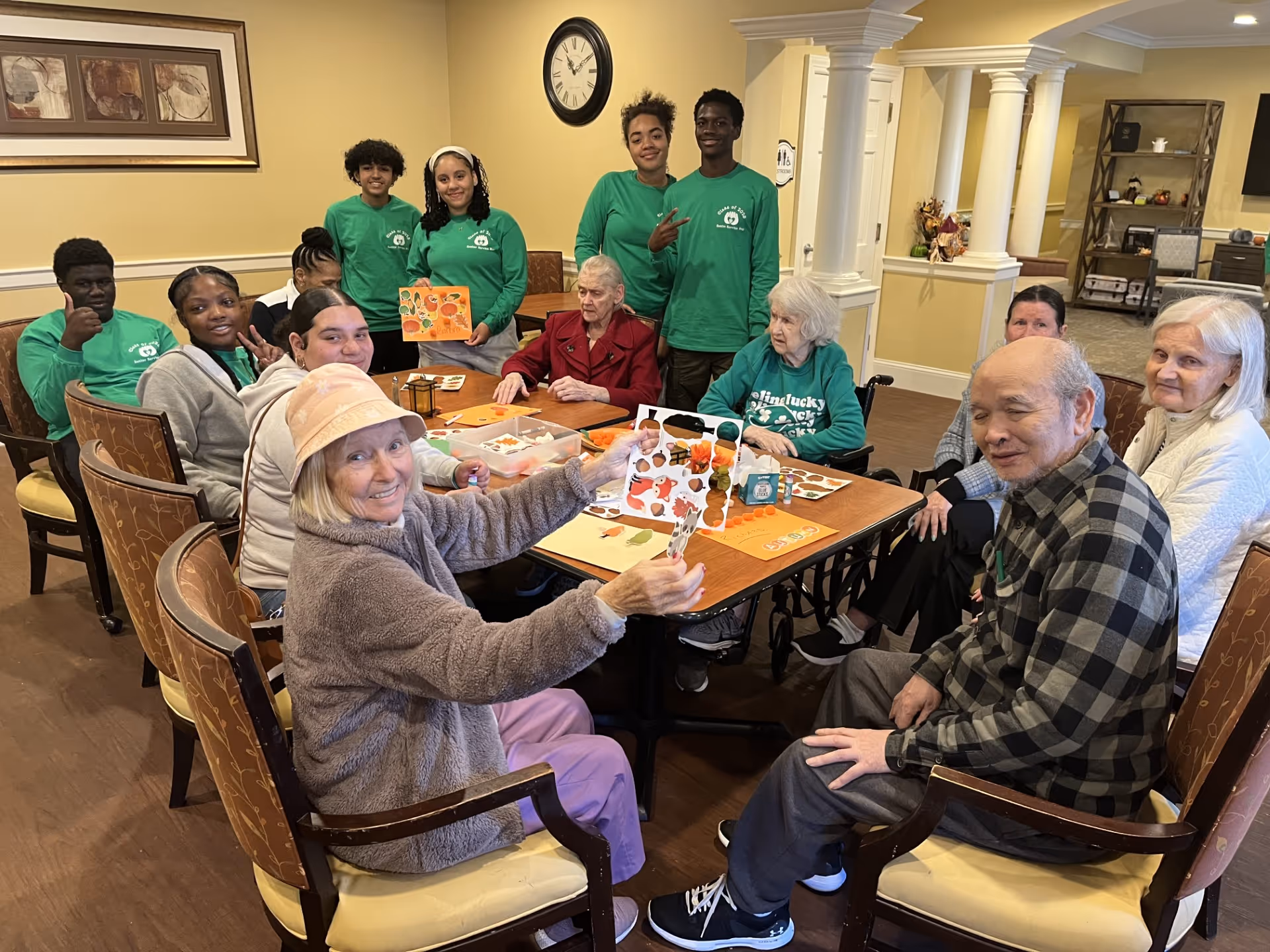 A group of elderly individuals and younger volunteers gathered around a table in a common room, engaging in a craft activity with paper cutouts and decorations. The room has warm yellow walls, a clock, framed artwork, and white columns in the background.