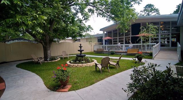 Outdoor garden area with a central black fountain surrounded by a circular flower bed. Four wooden chairs are placed around the fountain on a grassy patch. A large tree provides shade, and there is a paved walkway curving around the garden. In the background, there is a building with a screened porch and a ramp leading up to it.