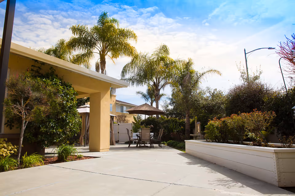 Outdoor patio area at Carlton Senior Living San Jose with palm trees, shrubs, and a seating area featuring a table and chairs under a large umbrella. The sky is partly cloudy and the area is surrounded by greenery and a beige building structure.