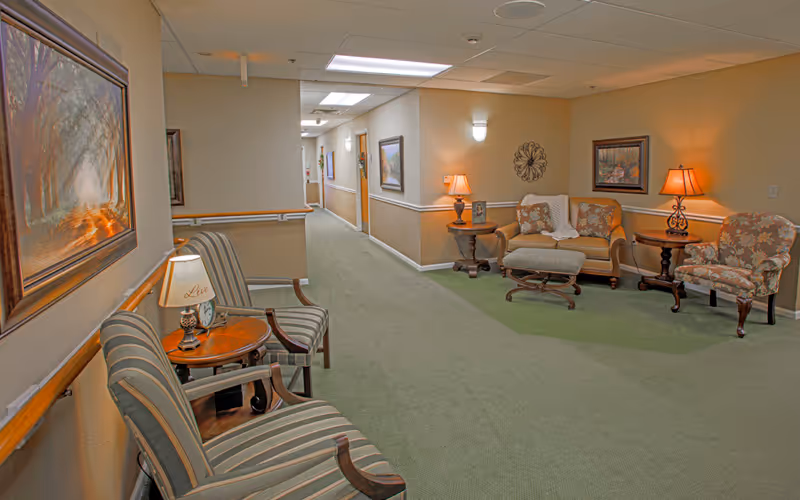 A well-lit hallway in a senior living facility with green carpet and beige walls. On the left side, there are two striped armchairs with a small round wooden table and a lamp between them. On the right side, there is a seating area with a loveseat, an ottoman, a floral armchair, two side tables with lamps, and framed artwork on the walls. The hallway extends into the distance with doors and more artwork visible.