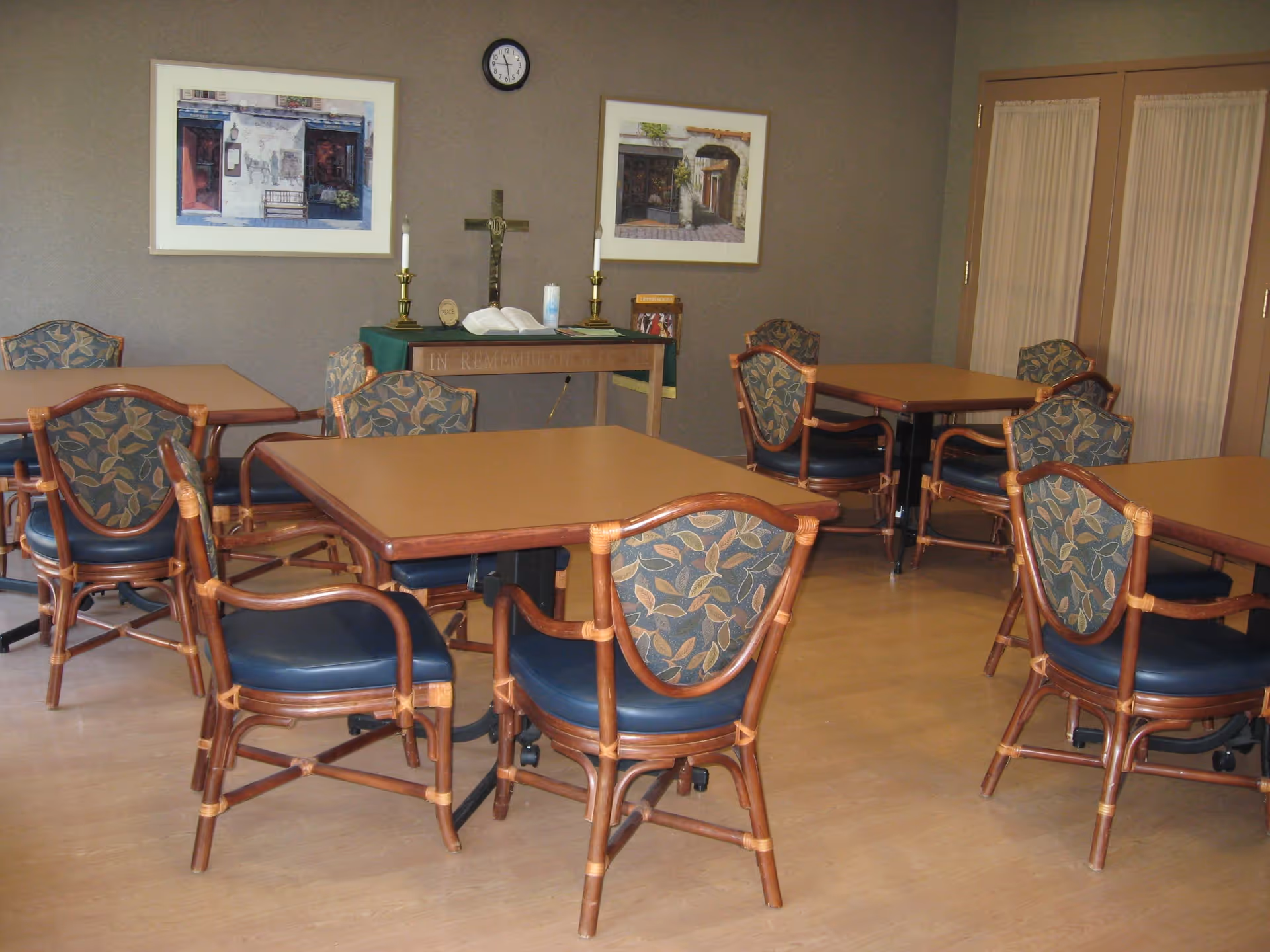 A quiet room with several square tables and wooden chairs with blue cushioned seats and leaf-patterned backs. On the back wall, there is a small altar with a cross, two candles, an open book, and a small sign. Two framed pictures hang on the wall above the altar, and a clock shows the time as 11:10. The room has wooden flooring and beige walls with a set of double doors with curtains on the right side.
