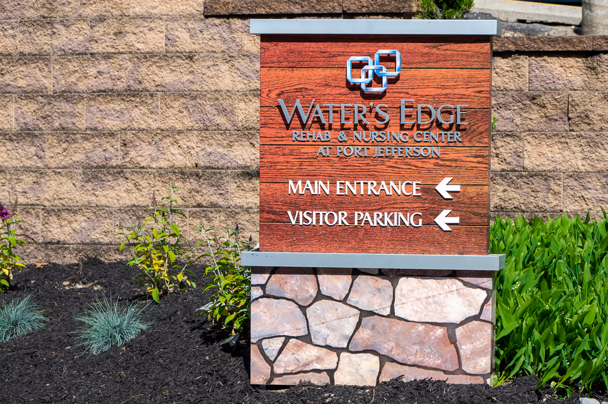 A sign for Water's Edge Rehab & Nursing Center at Port Jefferson with directions to the main entrance and visitor parking, set against a stone wall with plants and mulch around the base.