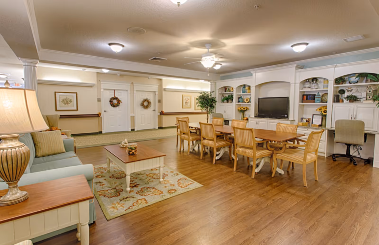A spacious senior living common area with wooden flooring, a light blue sofa with striped cushions, a wooden coffee table on a floral rug, a long wooden dining table with eight chairs, built-in white shelves and cabinets decorated with plants, books, and sunflowers, a flat-screen TV, and a small desk with an office chair. The walls are light-colored with framed artwork and two white doors adorned with wreaths.
