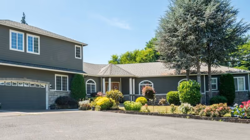 Exterior view of a single-story residential building with a two-car garage, surrounded by a well-maintained garden with various shrubs, flowers, and trees under a clear blue sky.