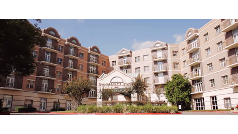 Exterior front view of University Place Senior Living Community, a multi-story residential building with balconies, trees, and landscaping under a blue sky.