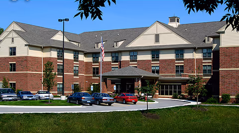 Exterior view of a multi-story senior living facility building with a brick and beige facade, a covered entrance, several parked cars in front, and a flagpole with an American flag.