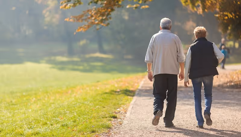 Two elderly people walking on a gravel path in a park-like outdoor setting with green grass and trees with autumn-colored leaves.