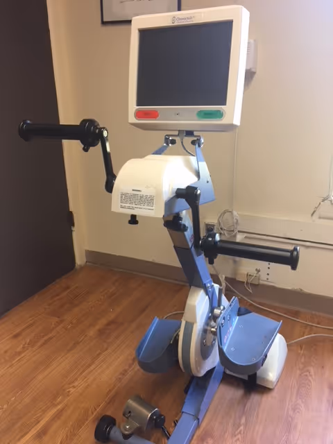A medical exercise machine with handles and foot pedals, equipped with a digital screen, placed on a wooden floor against a beige wall in an indoor setting.