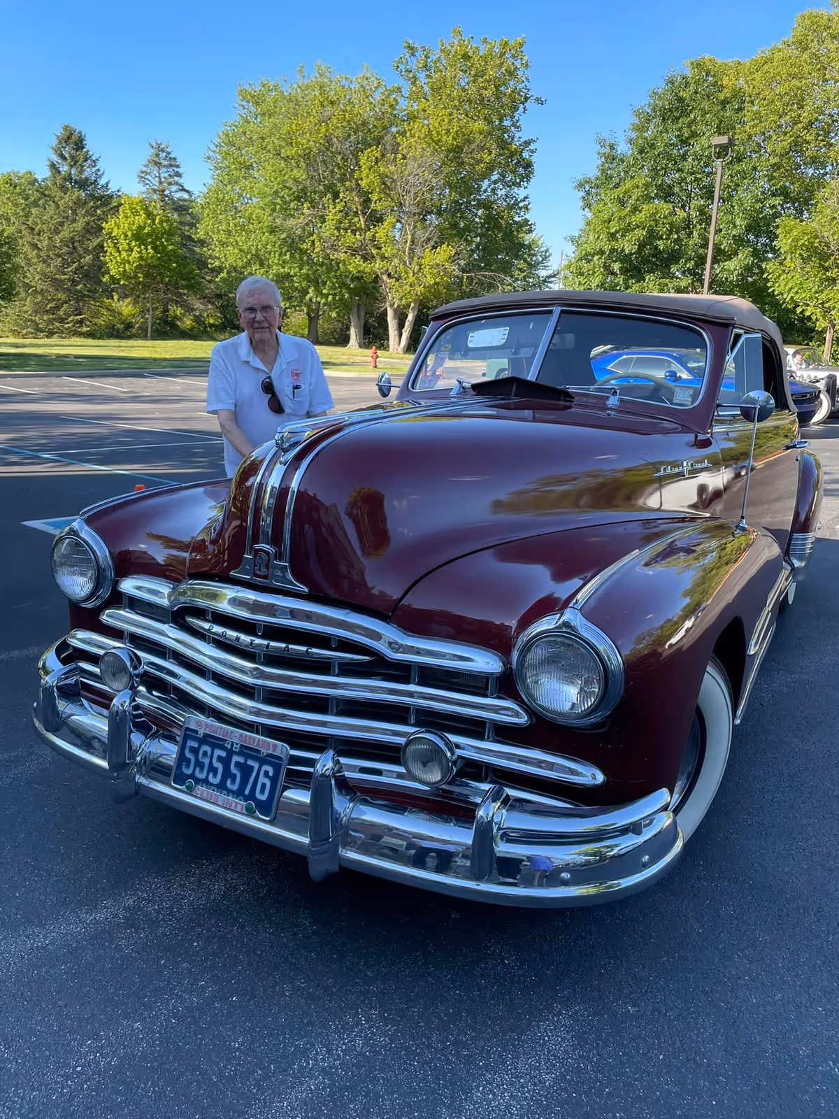 An elderly man wearing a white shirt stands behind a shiny maroon vintage Pontiac convertible car parked in an outdoor parking lot with green trees and a clear blue sky in the background.