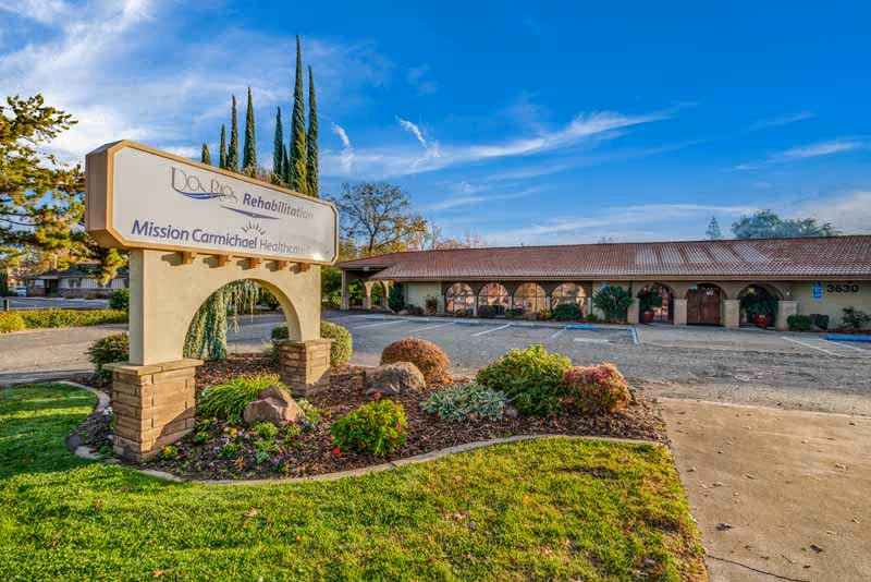 Exterior view of Mission Carmichael HealthCare Center showing a single-story building with arched windows and a tiled roof. In front of the building is a landscaped area with bushes and rocks, and a large sign that reads 'Do Reo Rehabilitation Mission Carmichael HealthCare'. The sky is clear and blue.