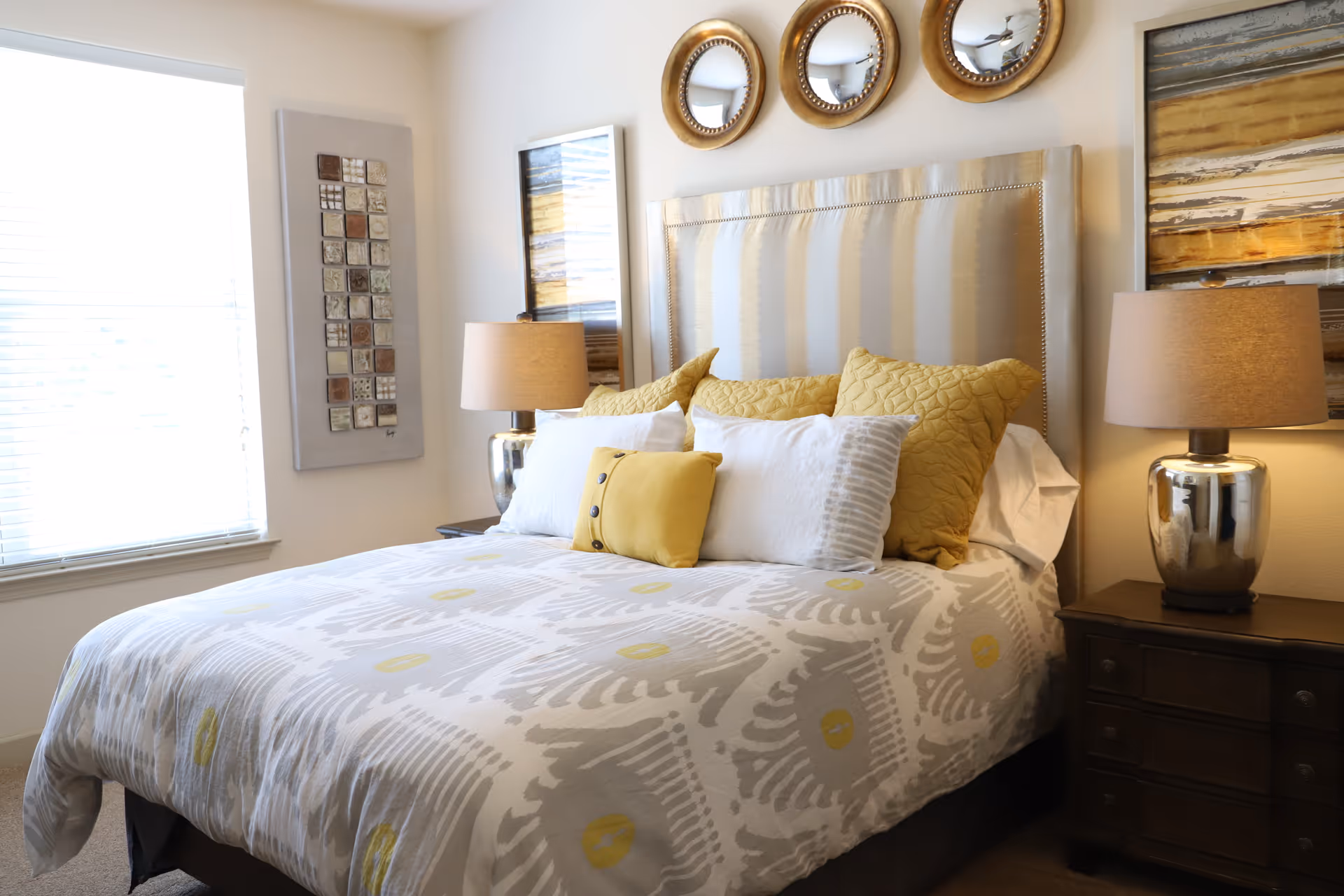 A neatly made bed with a patterned white and gray bedspread featuring yellow accents, several pillows in white and yellow, and a beige upholstered headboard. On either side of the bed are dark wooden nightstands with matching silver lamps with beige lampshades. Above the headboard are three round decorative mirrors, and there are framed artworks on the walls. A window with blinds is visible on the left side of the room.