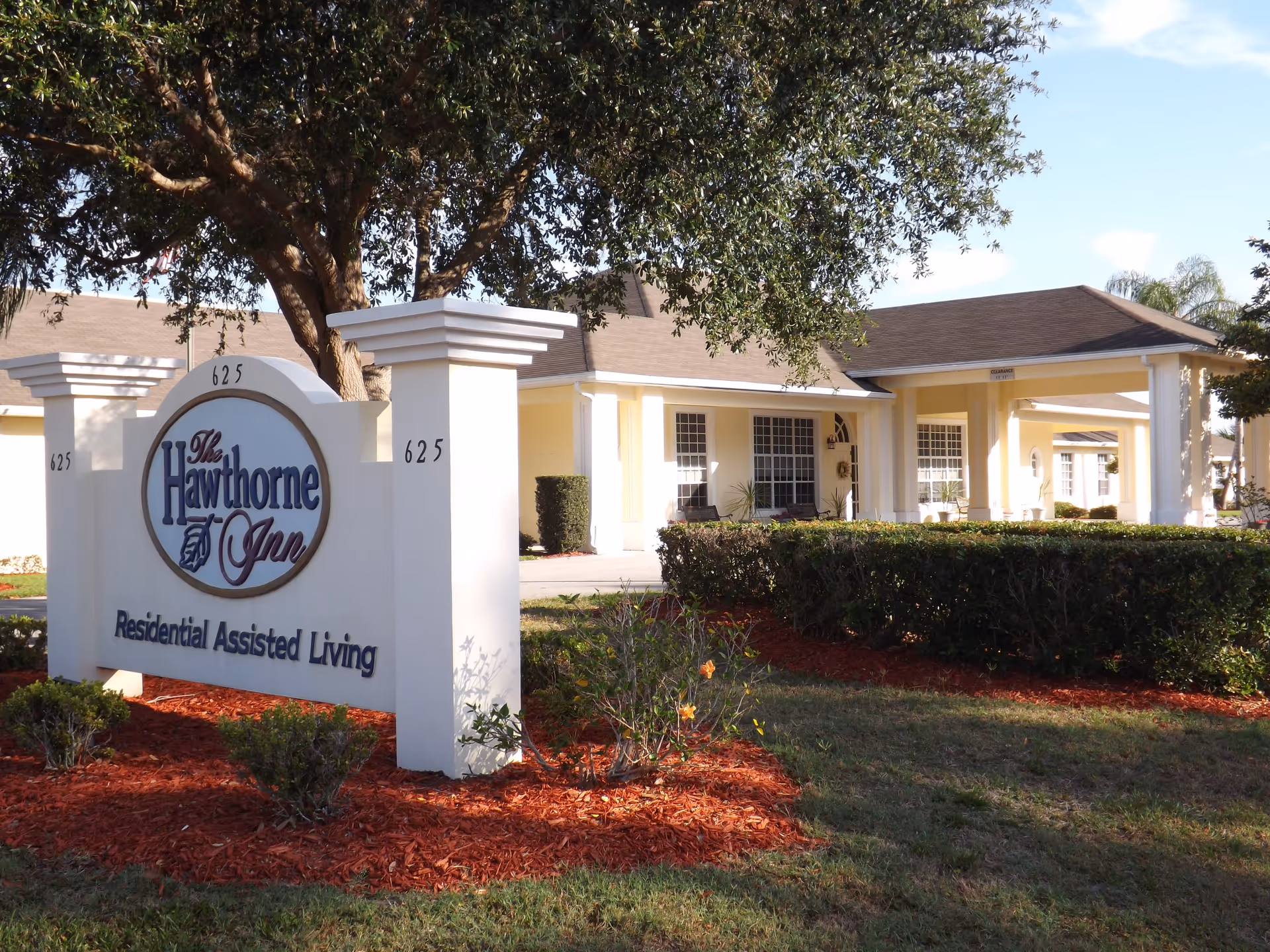 Exterior view of Hawthorne Inn Residential Assisted Living facility with a large sign displaying the facility name and address number 625, surrounded by landscaping including bushes, a tree, and red mulch.