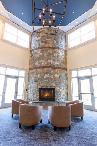 A cozy seating area with four beige armchairs arranged in a semicircle around a stone fireplace with a fire burning inside. The room has large windows letting in natural light, a high ceiling painted blue, and a modern chandelier hanging above.