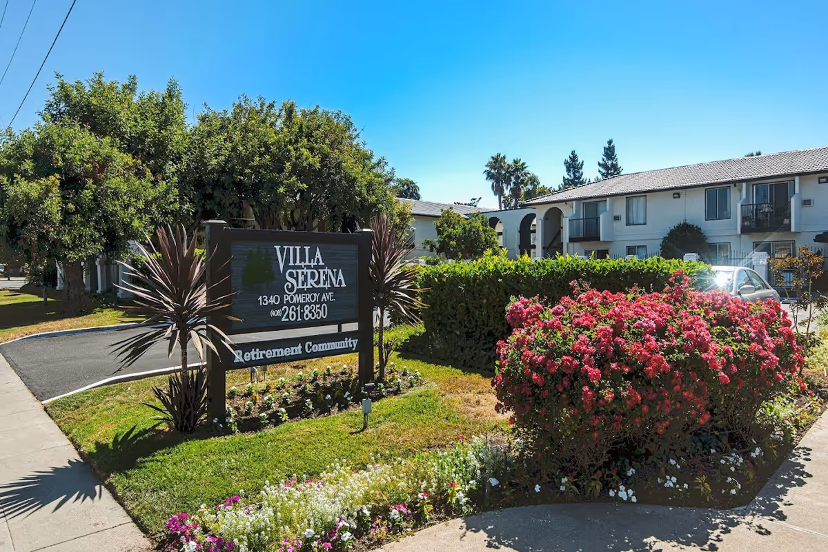 Exterior view of Villa Serena Retirement Community with a sign displaying the name, address, and phone number. The area is landscaped with green bushes, colorful flowers, and trees under a clear blue sky. A two-story building with balconies is visible in the background.