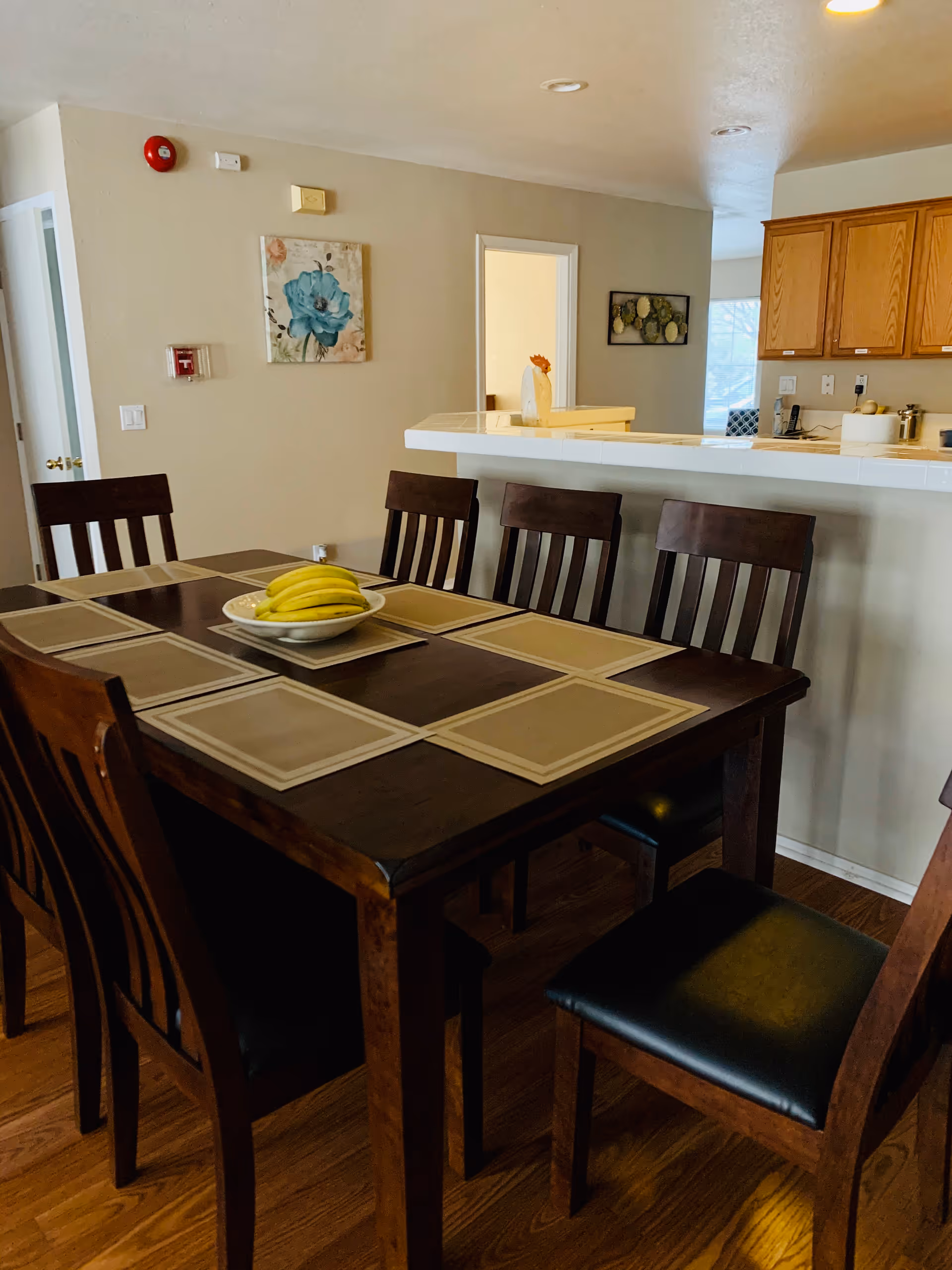 Dining area with a dark wooden table set with eight chairs, each with a beige placemat. A bowl of bananas is placed in the center of the table. In the background, there is a kitchen counter with wooden cabinets and a few kitchen items. The walls are light-colored with two framed pictures hanging.