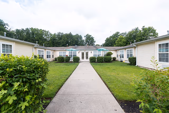 A single-story senior living facility building with beige siding, surrounded by green grass and bushes. A concrete walkway leads to the entrance with double glass doors. Two blue umbrellas provide shade over outdoor seating areas near the entrance. Trees are visible in the background under a cloudy sky.