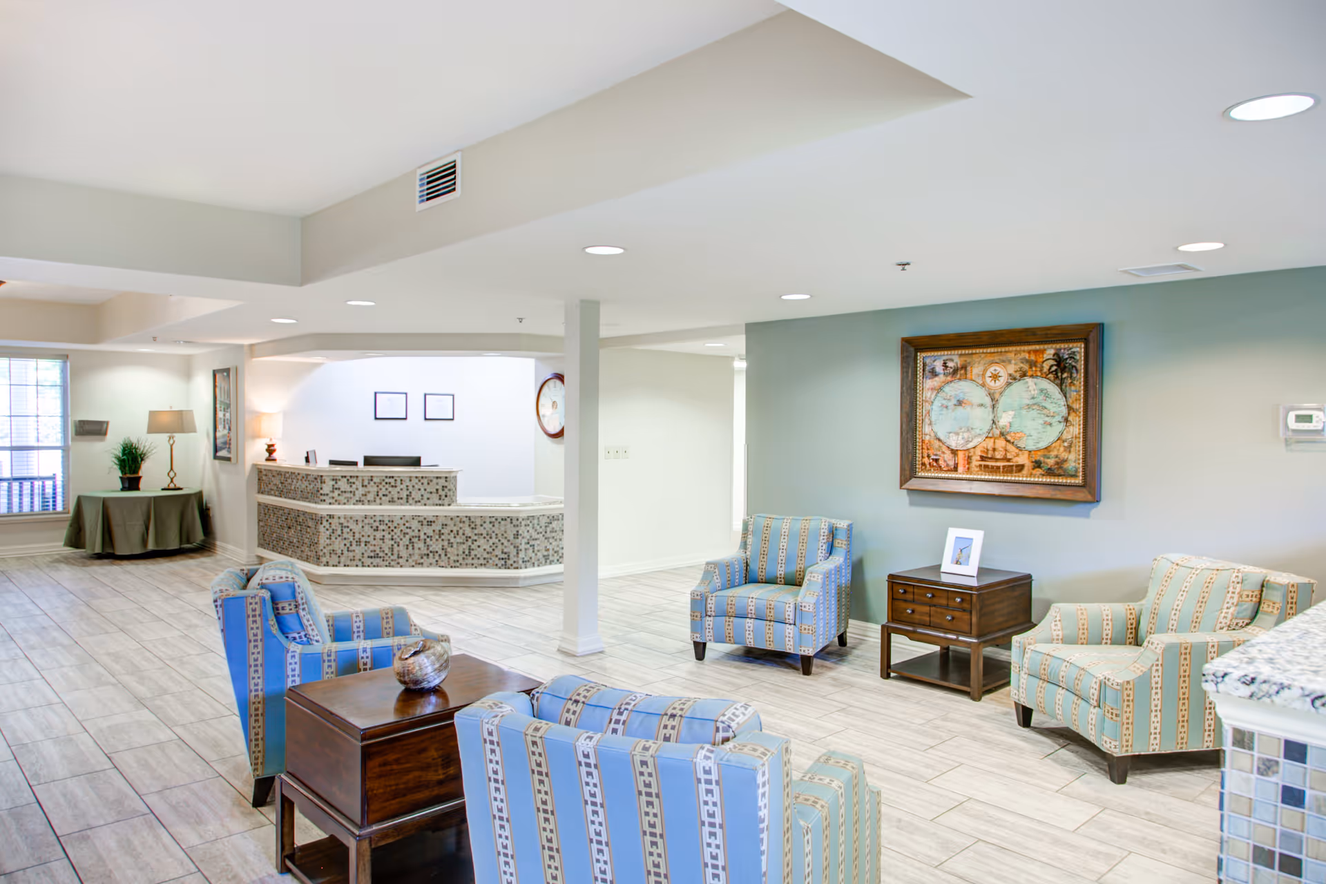 A bright and spacious senior living facility lobby with a reception desk in the background. The seating area includes several patterned armchairs in blue and beige tones arranged around wooden side tables. The walls are painted light colors, with a framed world map artwork hanging on one wall. The floor is covered with light-colored tiles, and there is a small table with a lamp and plant near a window.