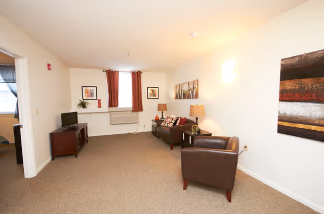 A living room area with beige carpet and white walls. There is a brown leather armchair and a dark brown sofa with patterned cushions. Two side tables with lamps are next to the sofa. A small TV sits on a wooden stand near the left wall. The window has red curtains, and there are framed pictures and a plant on the window sill. A colorful abstract painting hangs on the right wall.