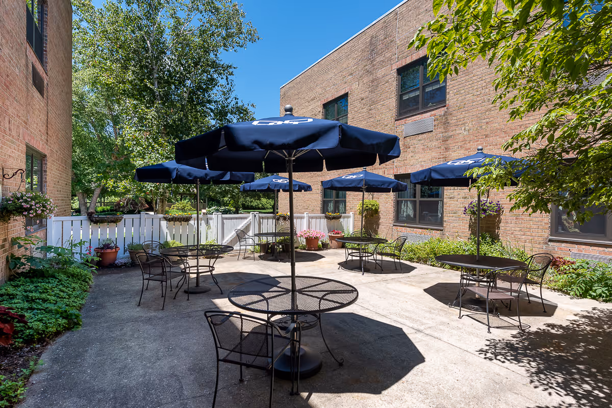 Outdoor patio area with several round metal tables and chairs, each table shaded by a blue umbrella. The patio is surrounded by brick building walls, white fencing, and greenery including trees and potted plants under a clear blue sky.