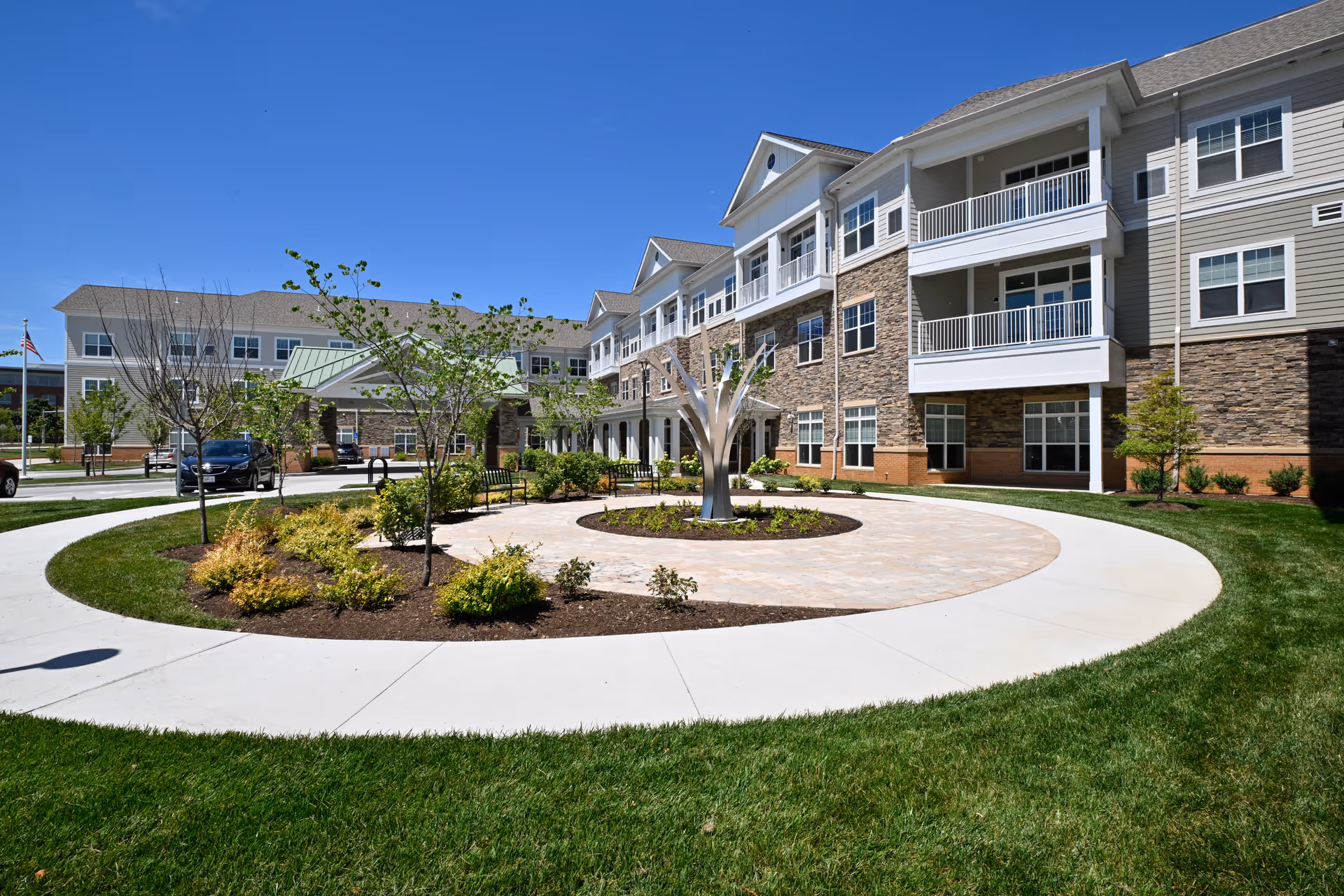 Exterior view of The Lumiere of Chesterfield senior living facility on a clear sunny day, showing a three-story building with balconies, stone and siding facade, landscaped circular garden with a modern metal sculpture in the center, and a paved walkway surrounded by green grass.