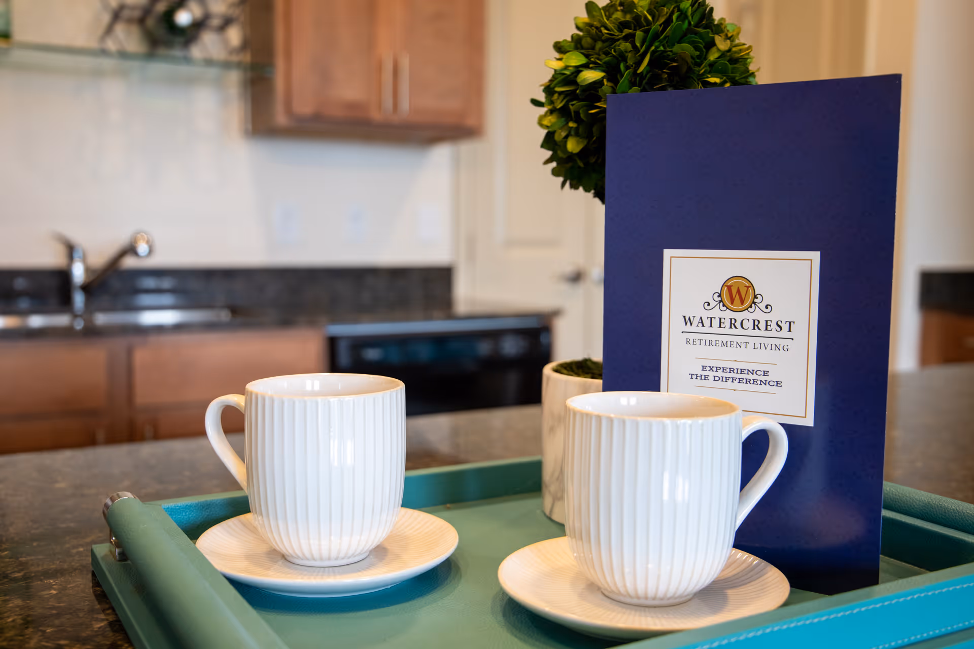 Two white ceramic coffee cups on matching saucers placed on a teal tray on a kitchen countertop. Behind the cups is a small potted plant and a blue brochure with the Watercrest Retirement Living logo and the text 'Experience the Difference'. The background shows a kitchen sink, wooden cabinets, and a dishwasher.