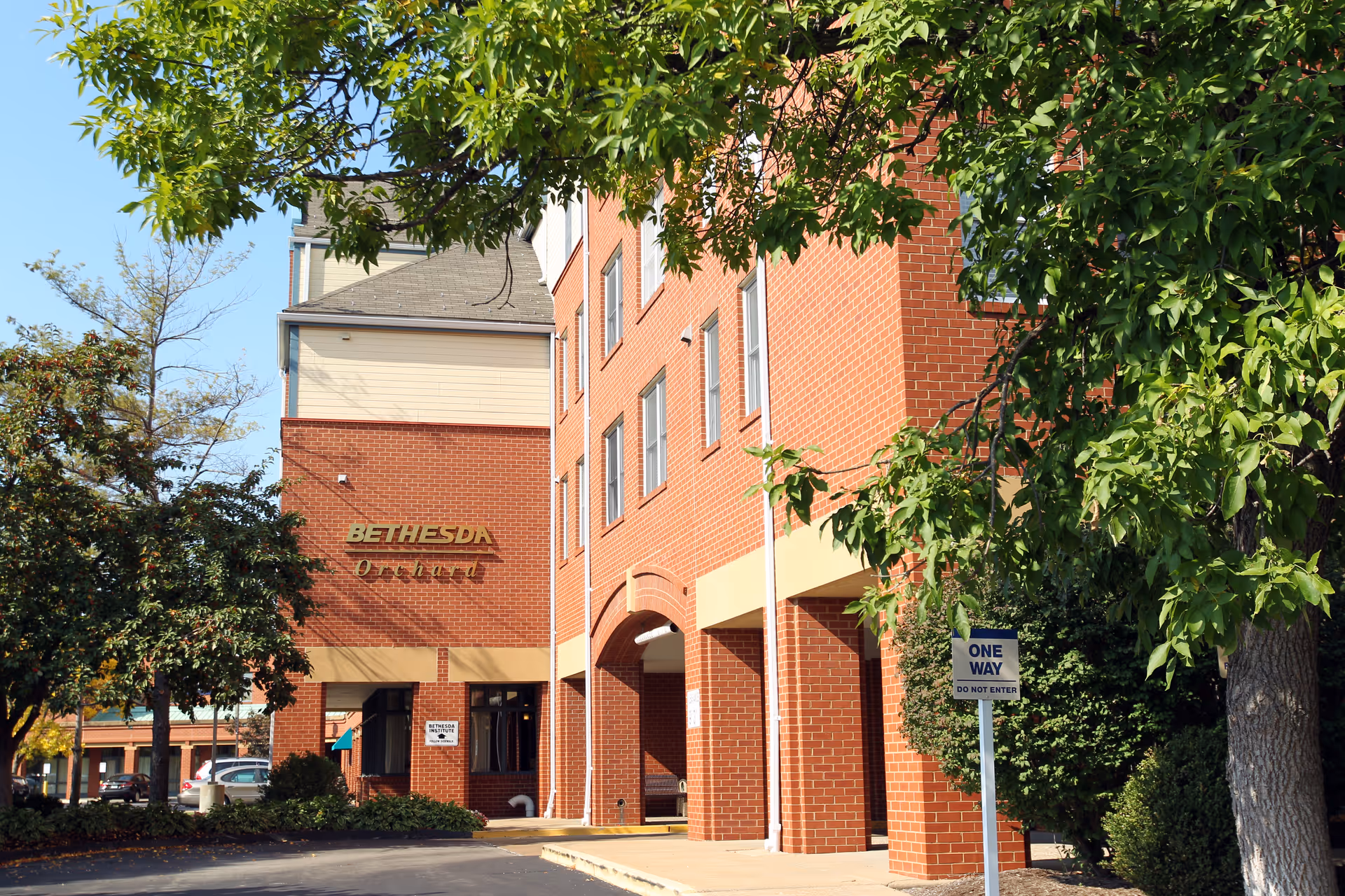 Exterior view of a brick building with the sign 'Bethesda Orchard' on the wall. The building is surrounded by trees and bushes, and there is a 'One Way Do Not Enter' sign visible near the sidewalk.