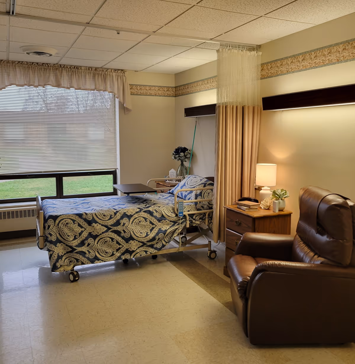 A nursing facility room with a hospital bed covered in a blue and beige patterned blanket, a bedside table with a lamp and decorative items, a brown leather recliner chair, and a large window with blinds and a valance. There is a privacy curtain partially drawn around the bed.