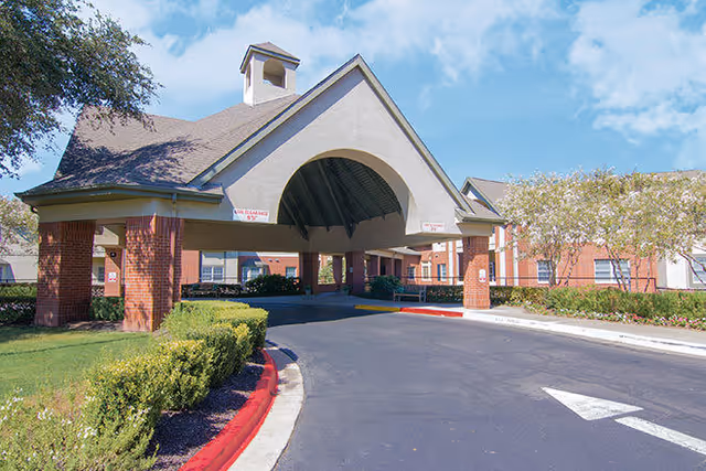 Entrance driveway and covered drop-off area of a senior living facility with brick pillars, manicured bushes, and a clear blue sky.