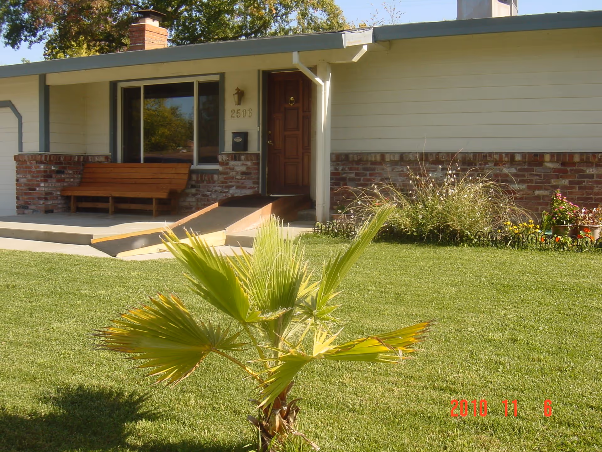 Front exterior view of a single-story house with a brick and siding facade, a wooden bench on the porch, a ramp leading to the front door, and a small palm tree in the grassy front yard.