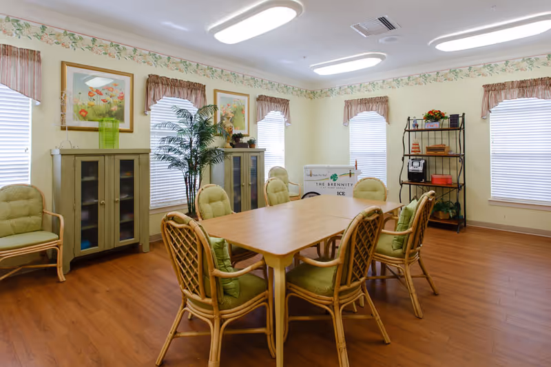 A bright and cozy dining room with a wooden table surrounded by eight cushioned chairs with green upholstery. The room has light yellow walls with floral border wallpaper near the ceiling, three windows with white blinds and pink valances, two green cabinets with glass doors, a potted plant, framed floral artwork, and a black metal shelf holding books and decorative items. There is also an ice cart labeled 'The Brennity' in the corner.
