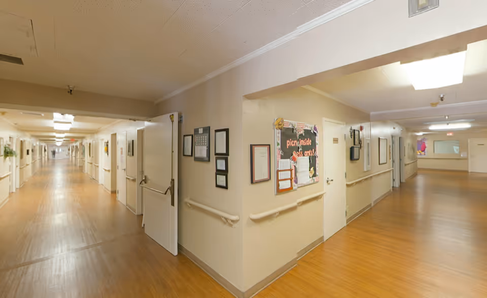 Interior view of a long, well-lit hallway in a senior living facility with light-colored walls, wooden flooring, handrails along the walls, bulletin boards, and multiple closed doors on both sides.