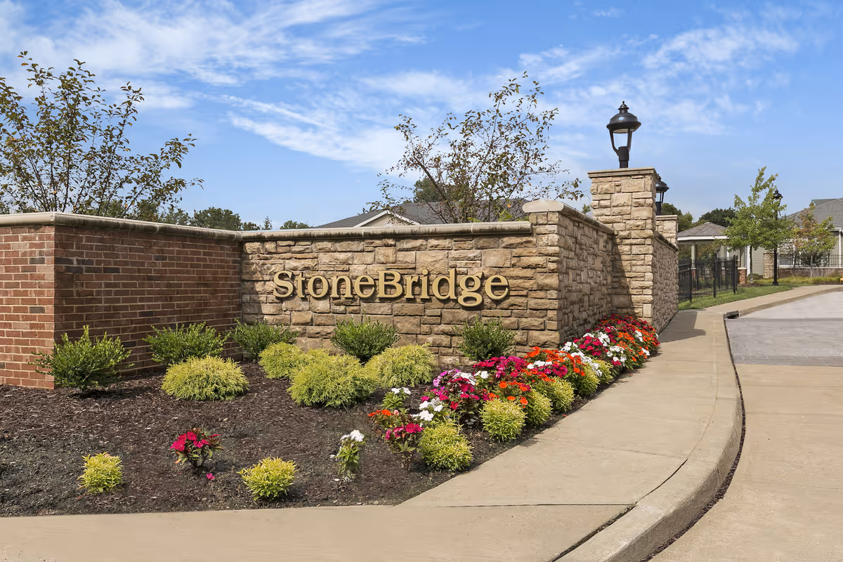StoneBridge Senior Living entrance sign made of stone with gold lettering, surrounded by landscaped bushes and colorful flowers, with a clear blue sky in the background.