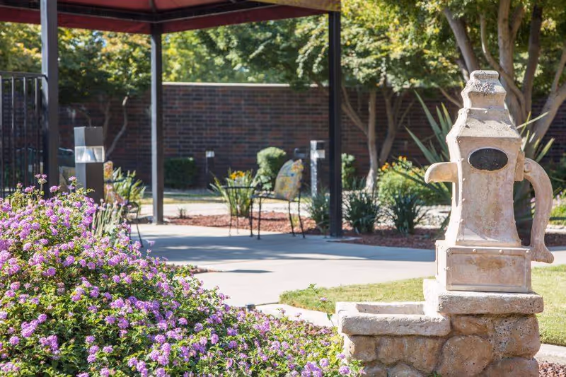Outdoor garden area with a stone water fountain in the foreground, purple flowering bushes, a covered patio with metal chairs and a brick wall in the background surrounded by trees and greenery.