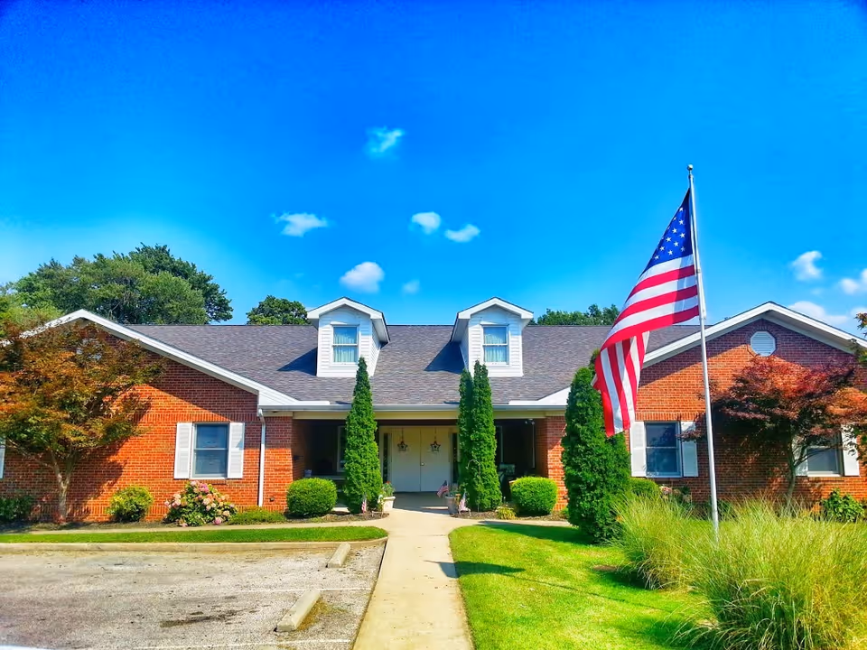 Front exterior view of a single-story brick building with a dark shingled roof and two dormer windows. There is a concrete walkway leading to double doors at the entrance, flanked by tall evergreen shrubs. An American flag on a flagpole is prominently displayed on the right side of the walkway. The building is surrounded by well-maintained grass, bushes, and small trees under a bright blue sky with a few clouds.