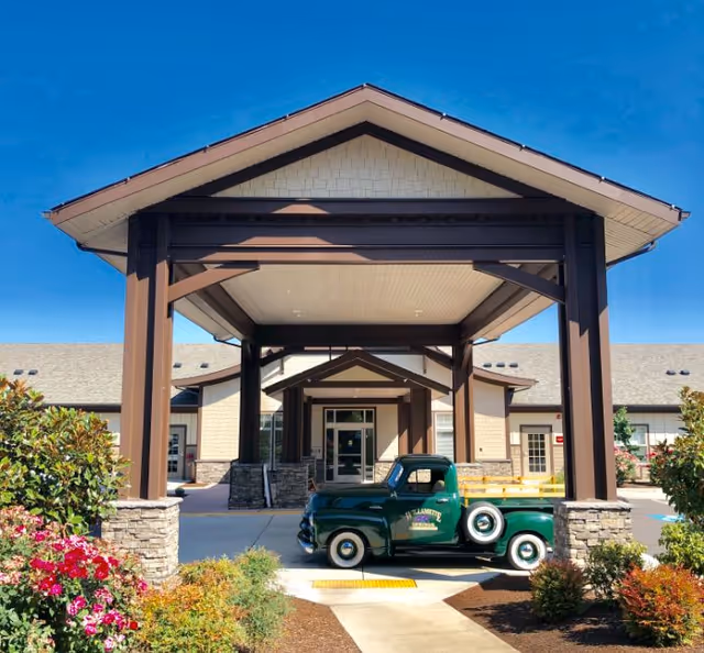 Entrance of Willamette Springs Memory Care Community featuring a covered driveway with wooden beams and stone pillars. A vintage green pickup truck with the facility's name on the door is parked under the covered area. Surrounding the entrance are landscaped bushes and flowers under a clear blue sky.