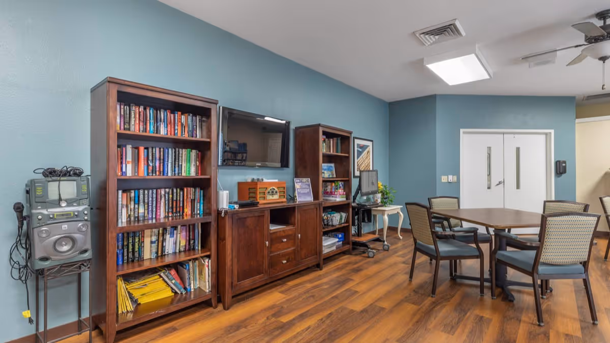 A cozy common area with wooden flooring and light blue walls. The room features two wooden bookshelves filled with books, a wooden cabinet with a flat-screen TV mounted above it, and a small table with a computer on a rolling cart. There is a square wooden table surrounded by six chairs with cushioned seats and backs. The ceiling has a fan and a rectangular light fixture, and there are double white doors in the background.