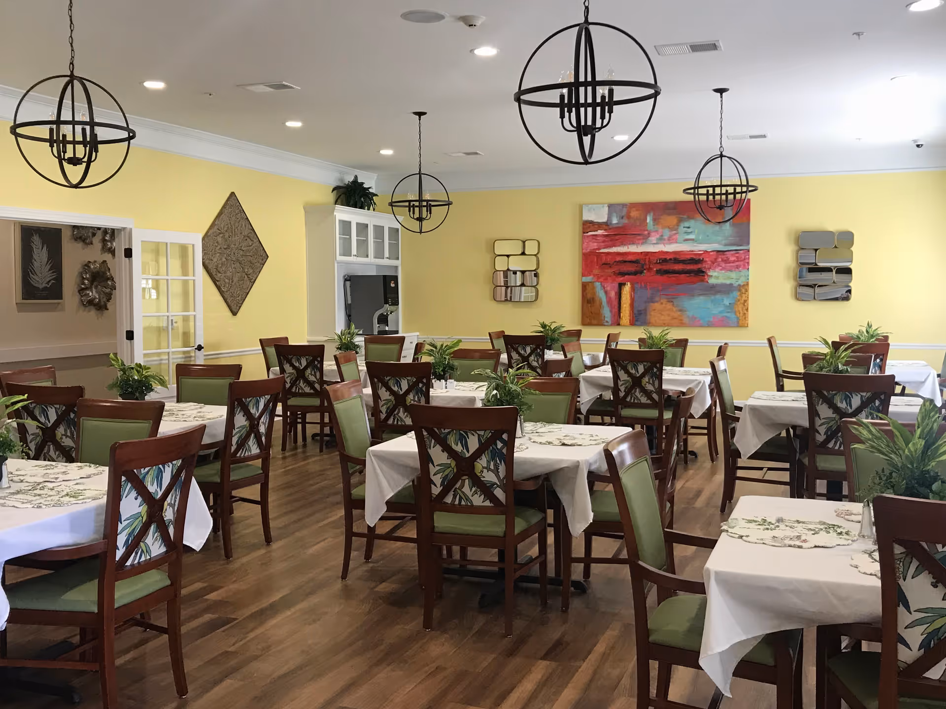 A dining room with multiple tables covered with white tablecloths and floral placemats. Each table is surrounded by wooden chairs with green cushions and floral patterned backs. The room has yellow walls decorated with colorful abstract paintings and mirrors. There are three spherical black chandeliers hanging from the ceiling, and several potted plants are placed on the tables. The floor is wooden, and there is a doorway with glass-paneled double doors on the left side.