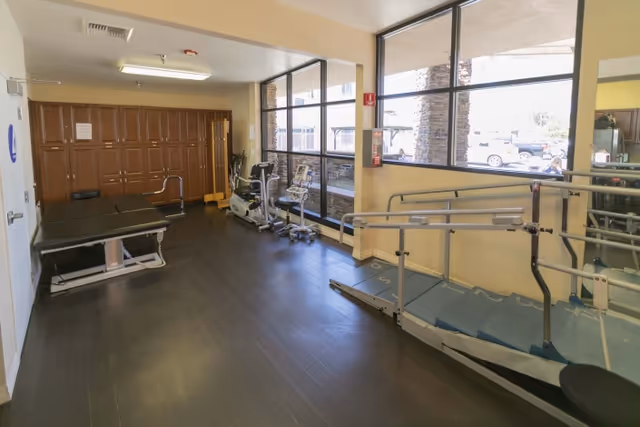 Interior room with exercise equipment including a treadmill with handrails, stationary bikes, and a padded examination or therapy table. The room has large windows letting in natural light and wooden cabinets along one wall.