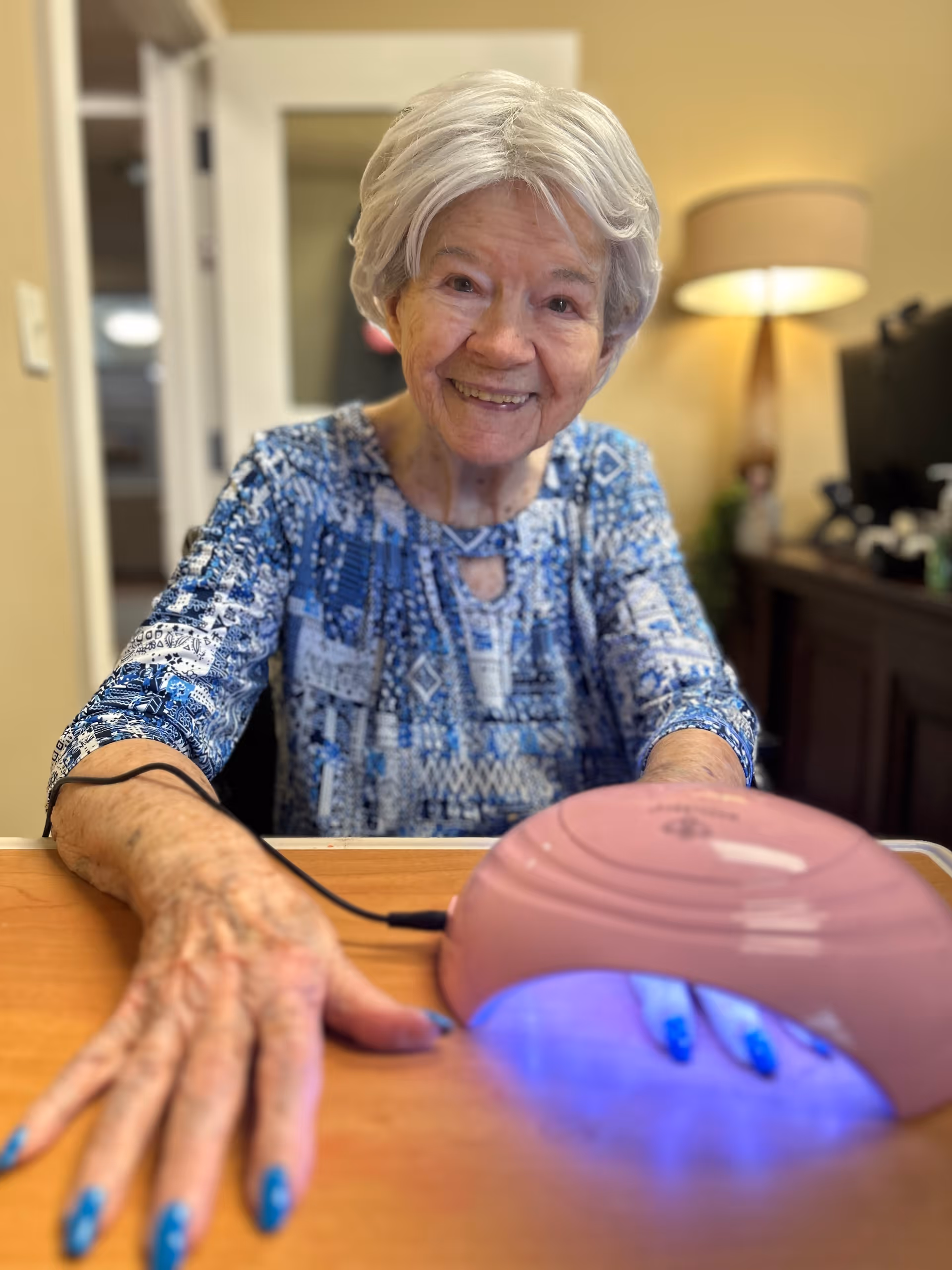 An elderly woman with short gray hair and a blue patterned blouse is smiling at the camera while having her nails done under a pink UV nail lamp emitting blue light. She is seated at a wooden table in a warmly lit room with a lamp and some furniture in the background.