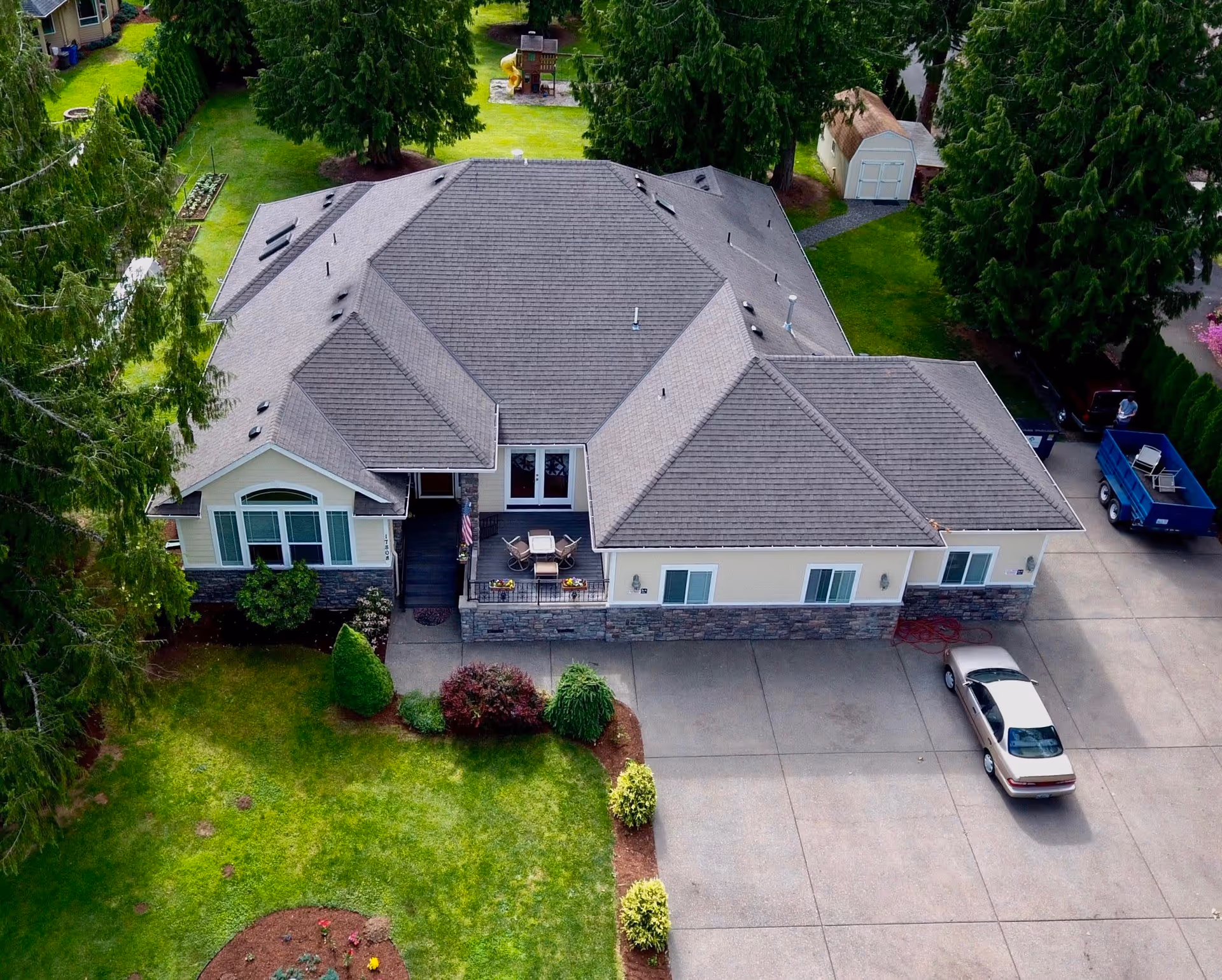 Aerial view of a single-story residential building with a gray roof, surrounded by green trees and a well-maintained lawn. There is a concrete driveway with a parked car and a blue trailer. The backyard has a small play structure and a shed.