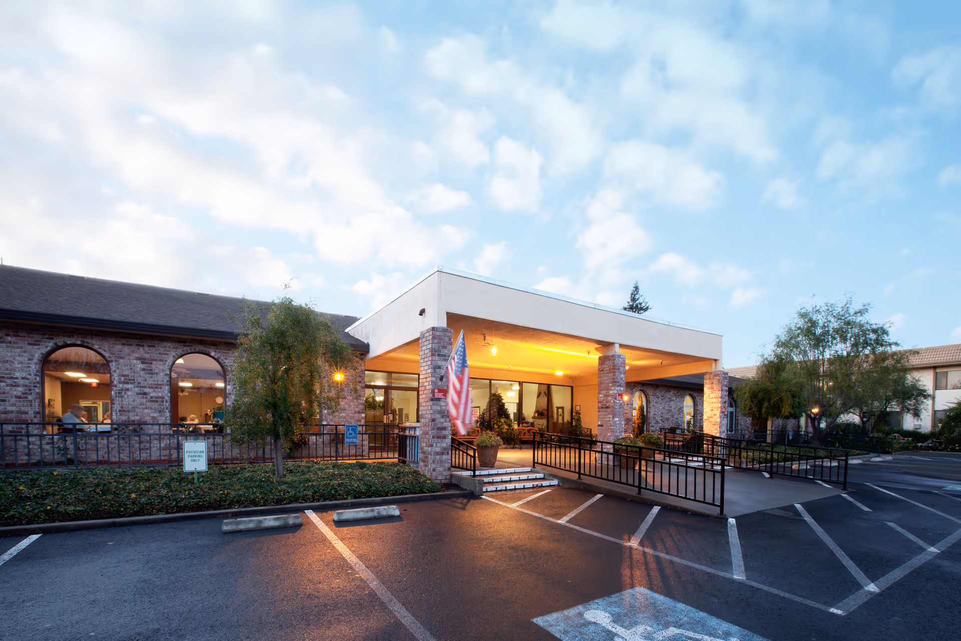 Exterior view of Napa Valley Care Center at dusk showing the entrance with a covered drop-off area supported by brick columns, an American flag, and a parking lot with designated handicapped parking spaces. The building has large arched windows and surrounding greenery.