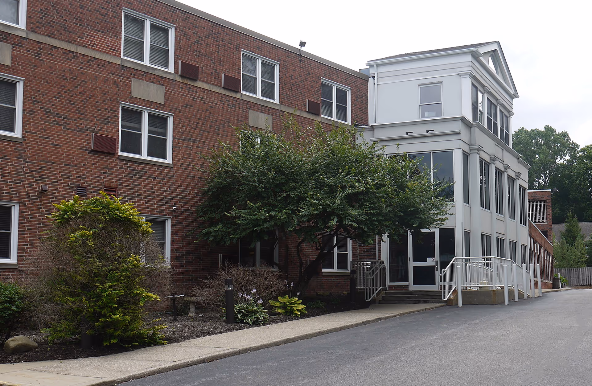 Exterior view of Cedarwood Plaza, a multi-story brick and white building with several windows, a small tree, bushes, and a paved driveway leading to an entrance with a ramp and stairs.