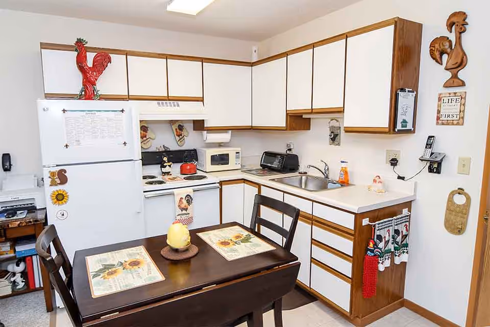 A small kitchen with white cabinets and wooden trim, featuring a white refrigerator, stove with a red kettle, microwave, toaster, and a sink. A dark wooden table with two chairs is set with sunflower-themed placemats and a decorative centerpiece. The kitchen is decorated with rooster-themed items and wall hangings.