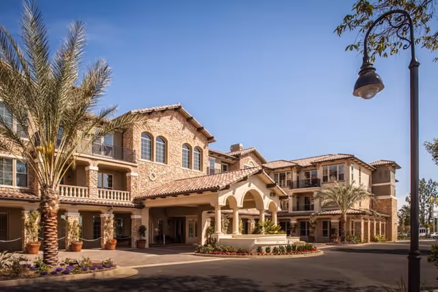 Exterior view of a multi-story retirement living facility with a Mediterranean architectural style, featuring stone and stucco walls, arched windows, balconies, palm trees, and a covered entrance driveway under a clear blue sky.