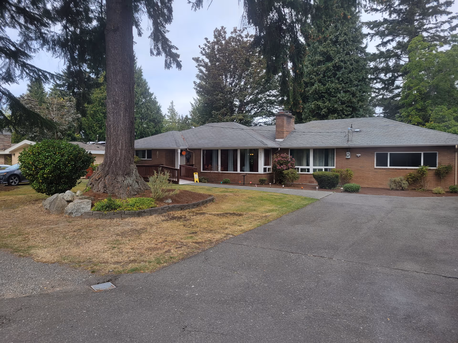Single-story brick building with a gray roof surrounded by trees and shrubs. There is a large tree in the front yard with a small landscaped area around its base. A paved driveway leads up to the building entrance, which has a small ramp and railing. The sky is overcast.