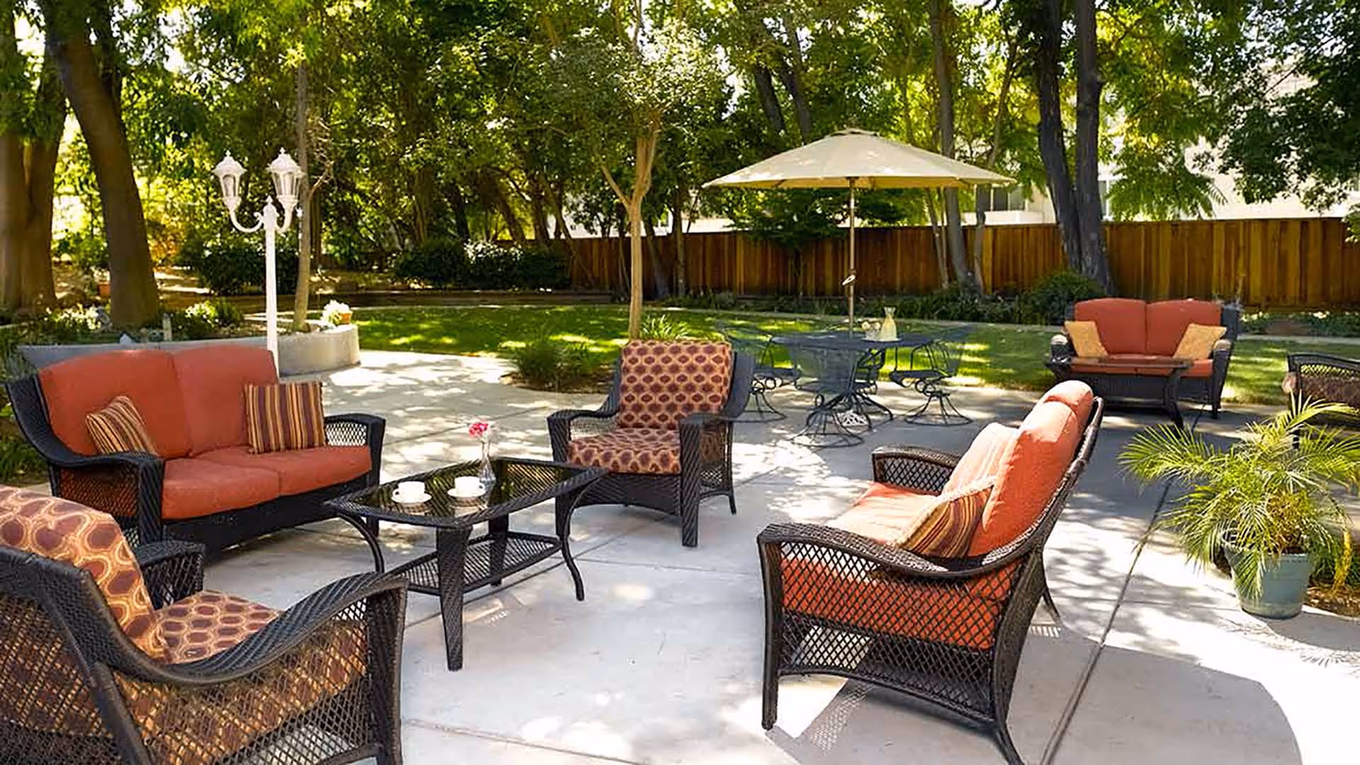 Outdoor patio area with wicker furniture including cushioned chairs and loveseats arranged around a glass coffee table. There is a round metal table with chairs and a large umbrella in the background. The patio is surrounded by green grass, trees, and a wooden fence.