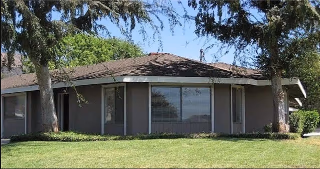 Single-story building with a brown roof and large windows, surrounded by green grass and trees under a clear blue sky.