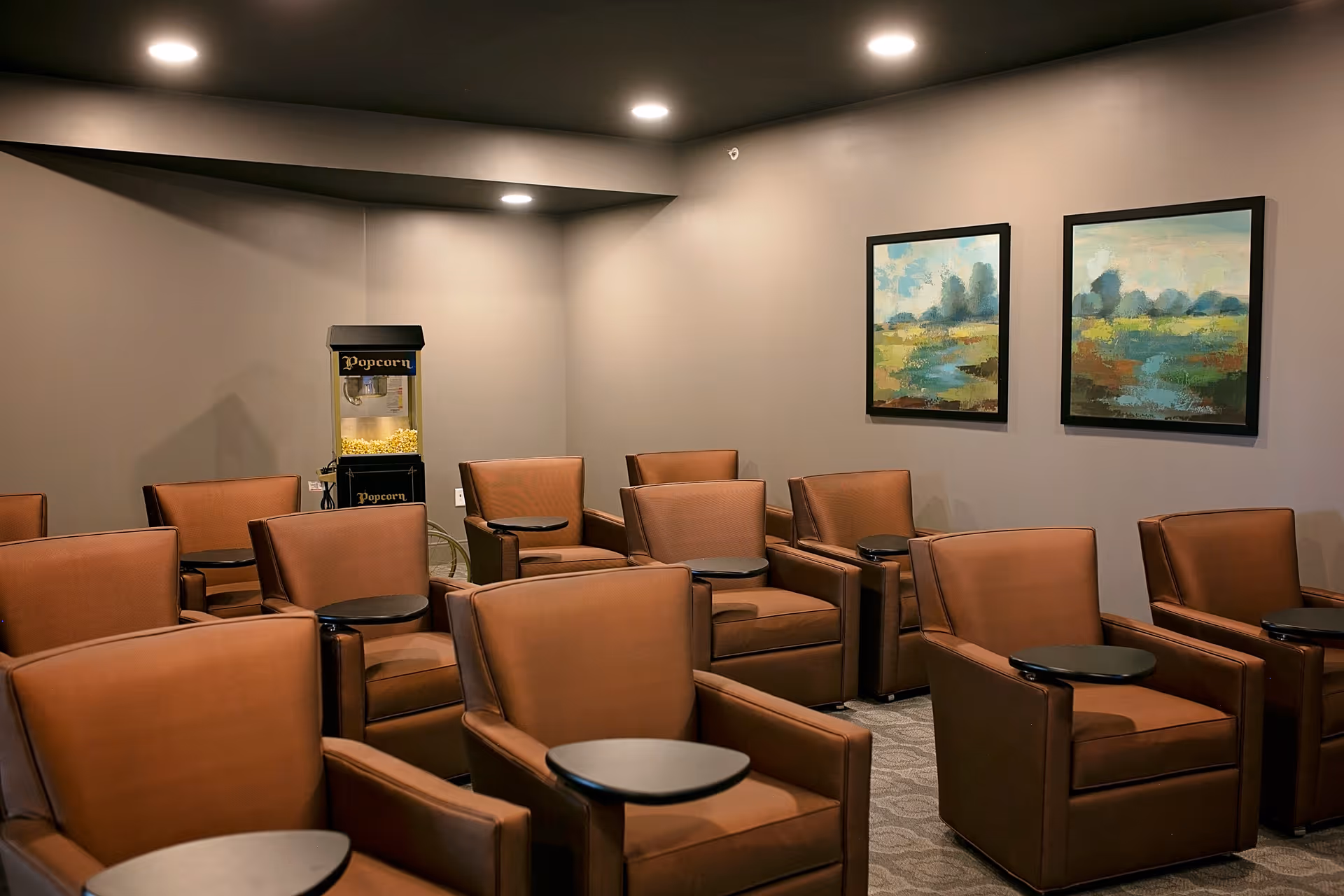 A small theater room with multiple brown leather armchairs, each equipped with a small black side table. There is a popcorn machine in the corner and two landscape paintings on the wall. The room has soft lighting and neutral-colored walls.
