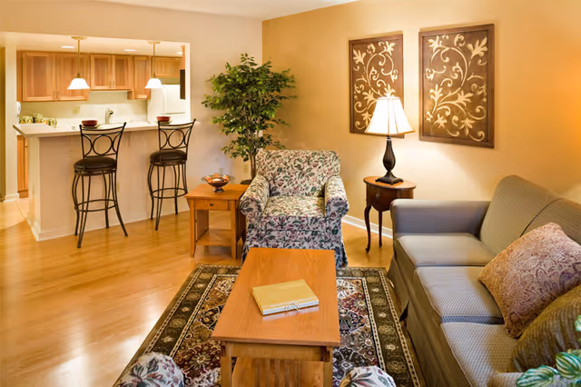 A cozy living room with a floral armchair, a gray sofa with patterned pillows, a wooden coffee table with a book on it, and a side table with a lamp. The room has warm lighting, two decorative wall hangings, and a green potted plant. In the background, there is a kitchen area with wooden cabinets, a white refrigerator, and two bar stools at a counter.